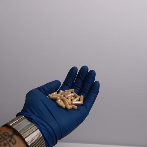 Hand wearing a blue glove holding gold-colored capsules against a gray background